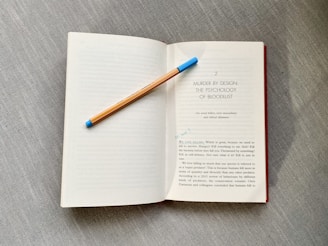 An open book rests on a textured gray surface. A pencil with a blue eraser lays diagonally across one of the pages. The book's content includes a title, 'Murder by Design: The Psychology of Bloodlust,' and a passage discussing topics related to killing and survival.