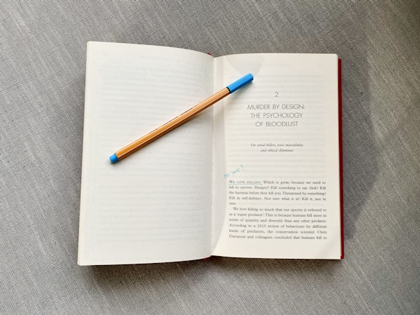 An open book rests on a textured gray surface. A pencil with a blue eraser lays diagonally across one of the pages. The book's content includes a title, 'Murder by Design: The Psychology of Bloodlust,' and a passage discussing topics related to killing and survival.
