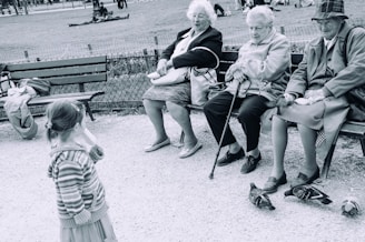 Three elderly women sit on a park bench, each wearing coats and holding personal items. They appear to engage with a young girl standing nearby, who wears a striped hooded sweater. Pigeons are on the ground near their feet. The setting is an outdoor park, with grass, trees, and more people visible in the background.