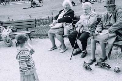 Three elderly women sit on a park bench, each wearing coats and holding personal items. They appear to engage with a young girl standing nearby, who wears a striped hooded sweater. Pigeons are on the ground near their feet. The setting is an outdoor park, with grass, trees, and more people visible in the background.