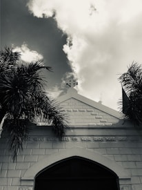A monochrome image featuring the facade of a building with a cross at its peak, flanked by palm trees. The sky is partly cloudy, adding depth and contrast to the scene.