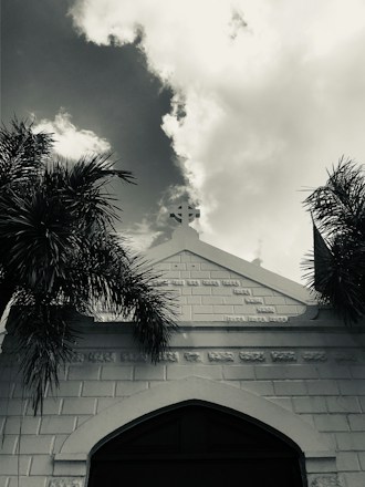 A monochrome image featuring the facade of a building with a cross at its peak, flanked by palm trees. The sky is partly cloudy, adding depth and contrast to the scene.