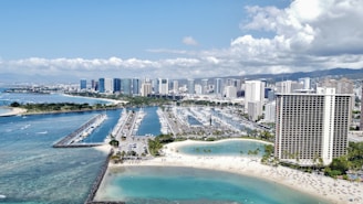 An aerial view of a coastal cityscape showcasing a marina filled with numerous boats, surrounded by turquoise ocean waters. High-rise buildings, including a prominent round hotel with balconies, dominate the background. Sandy beaches with scattered palm trees and people enjoying the sun are visible in the foreground, under a partly cloudy sky.