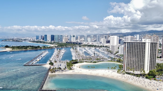 An aerial view of a coastal cityscape showcasing a marina filled with numerous boats, surrounded by turquoise ocean waters. High-rise buildings, including a prominent round hotel with balconies, dominate the background. Sandy beaches with scattered palm trees and people enjoying the sun are visible in the foreground, under a partly cloudy sky.