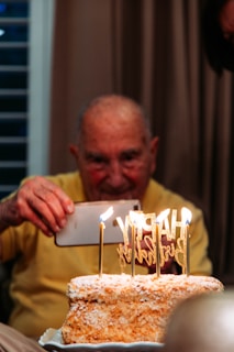 An elderly person in a yellow shirt is taking a photo of a rectangular birthday cake adorned with lit candles and a gold 'Happy Birthday' topper. The cake has an exterior covered in a crumbly texture, likely coconut or nuts, and is placed on a white decorative plate. The background features a softly lit, neutral curtain.