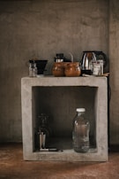 A rustic kitchen setup features a concrete shelf with various kitchen items. On the top, there are coffee mugs, a kettle, and some jars. Below, a glass water bottle, a metallic cup, and a corkscrew are placed along with a couple of glasses.