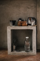 A rustic kitchen setup features a concrete shelf with various kitchen items. On the top, there are coffee mugs, a kettle, and some jars. Below, a glass water bottle, a metallic cup, and a corkscrew are placed along with a couple of glasses.