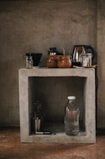 A rustic kitchen setup features a concrete shelf with various kitchen items. On the top, there are coffee mugs, a kettle, and some jars. Below, a glass water bottle, a metallic cup, and a corkscrew are placed along with a couple of glasses.