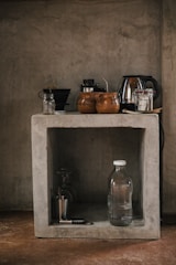 A rustic kitchen setup features a concrete shelf with various kitchen items. On the top, there are coffee mugs, a kettle, and some jars. Below, a glass water bottle, a metallic cup, and a corkscrew are placed along with a couple of glasses.