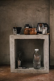 A rustic kitchen setup features a concrete shelf with various kitchen items. On the top, there are coffee mugs, a kettle, and some jars. Below, a glass water bottle, a metallic cup, and a corkscrew are placed along with a couple of glasses.