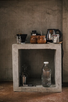 A rustic kitchen setup features a concrete shelf with various kitchen items. On the top, there are coffee mugs, a kettle, and some jars. Below, a glass water bottle, a metallic cup, and a corkscrew are placed along with a couple of glasses.