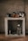 A rustic kitchen setup features a concrete shelf with various kitchen items. On the top, there are coffee mugs, a kettle, and some jars. Below, a glass water bottle, a metallic cup, and a corkscrew are placed along with a couple of glasses.