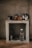 A rustic kitchen setup features a concrete shelf with various kitchen items. On the top, there are coffee mugs, a kettle, and some jars. Below, a glass water bottle, a metallic cup, and a corkscrew are placed along with a couple of glasses.