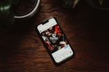 A smartphone lies on a wooden surface, displaying an Instagram post featuring a coffee shop logo on the screen. Surrounding the phone are blurred hints of potted plants.