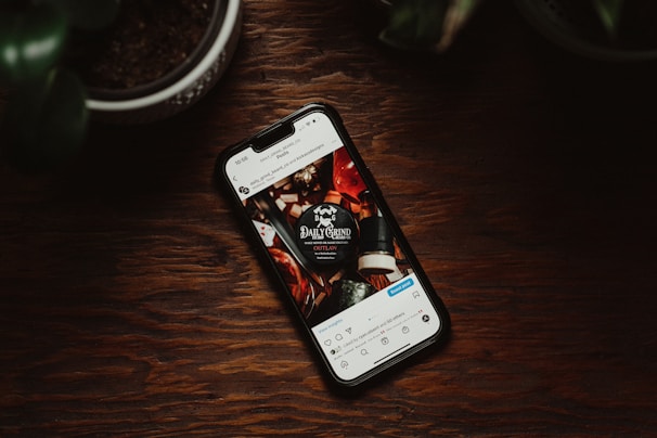 A smartphone lies on a wooden surface, displaying an Instagram post featuring a coffee shop logo on the screen. Surrounding the phone are blurred hints of potted plants.