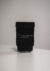 A black camera lens is standing upright on a light-colored surface with soft, diffused lighting in a minimalist setting. The focus is sharp on the lens, highlighting the engraved text and details on the lens body.