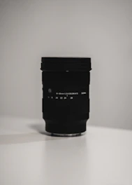 A sleek camera lens resting on a minimalist black surface with soft natural light highlighting its details.
