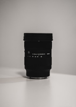 A black camera lens is standing upright on a light-colored surface with soft, diffused lighting in a minimalist setting. The focus is sharp on the lens, highlighting the engraved text and details on the lens body.