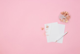 A flat lay composition featuring a pink background with two sheets of white paper, a white and gold pen, rose gold paper clips, and binder clips. A transparent dish on the upper right is filled with various paper clips and binder clips.