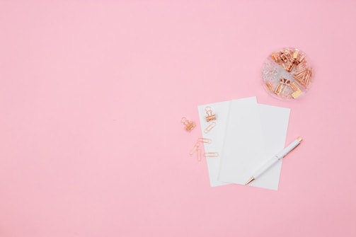 A flat lay composition featuring a pink background with two sheets of white paper, a white and gold pen, rose gold paper clips, and binder clips. A transparent dish on the upper right is filled with various paper clips and binder clips.