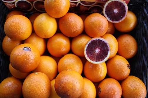 A basket filled with ripe oranges, with two oranges cut in half revealing their red interior. The oranges have a bright and fresh appearance, with some having small stickers on them.