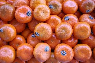 A pile of fresh, bright orange mandarins with stickers. Their smooth and glossy skins suggest they are ripe and ready to eat.