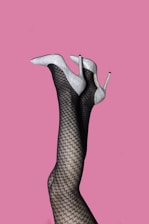 A pair of legs wearing houndstooth-patterned stockings are elevated against a plain pink background. The legs are adorned with sparkling silver high-heeled shoes.