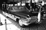 A vintage black car with chrome details is parked indoors. It features a classic design with a prominent front grille and round headlights. An Indonesian flag is attached to the front, and the surrounding area includes tiled flooring and hints of historical exhibits, including carriages in the background.