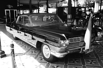 A vintage black car with chrome details is parked indoors. It features a classic design with a prominent front grille and round headlights. An Indonesian flag is attached to the front, and the surrounding area includes tiled flooring and hints of historical exhibits, including carriages in the background.