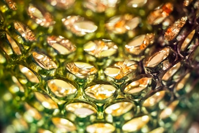 Close-up view of a textured surface resembling a honeycomb pattern, featuring translucent cells with a reflective, metallic sheen. The cells appear to be made of plastic or glass, capturing light in shades of green and gold, creating an intricate play of light and shadow.