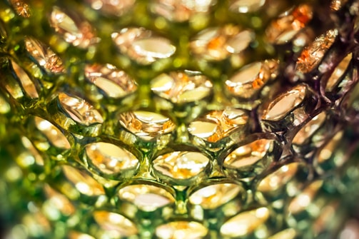 Close-up view of a textured surface resembling a honeycomb pattern, featuring translucent cells with a reflective, metallic sheen. The cells appear to be made of plastic or glass, capturing light in shades of green and gold, creating an intricate play of light and shadow.