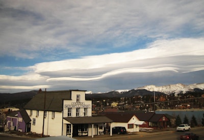 A small town scene with a prominent white building marked 'Fraser Mercantile Company' in the foreground. The building is surrounded by a few smaller houses and vehicles parked along the road. In the background, snow-capped mountains stretch across the horizon under a sky filled with unique, layered cloud formations.