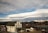 A small town scene with a prominent white building marked 'Fraser Mercantile Company' in the foreground. The building is surrounded by a few smaller houses and vehicles parked along the road. In the background, snow-capped mountains stretch across the horizon under a sky filled with unique, layered cloud formations.