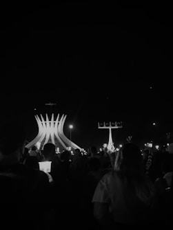 A crowd is gathered at night, facing a large, illuminated cathedral structure with tall, pointed arches. The scene is predominantly dark but illuminated by multiple small lights held by the people, creating a serene and contemplative atmosphere.