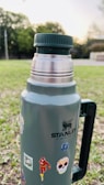 A metal thermos with a dark green cap and handle is placed outdoors on a grassy area. The thermos is adorned with various stickers, including images of superheroes and colorful designs. Trees and bushes are visible in the blurred background, suggesting a park or open area at sunset.