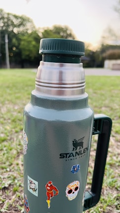 A metal thermos with a dark green cap and handle is placed outdoors on a grassy area. The thermos is adorned with various stickers, including images of superheroes and colorful designs. Trees and bushes are visible in the blurred background, suggesting a park or open area at sunset.