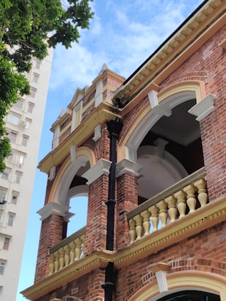 A historic brick building with intricate architectural details such as arched windows and ornate balustrades, partially framed by a leafy tree in the foreground. A modern high-rise building can be seen in the background under a clear blue sky.