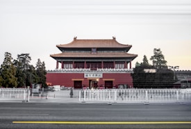 A grand traditional Chinese architectural building with multiple tiers and ornate roofing, surrounded by lush greenery and trees. A row of white barricades in front, and the building features intricate woodwork and a striking red fa&ccedil;ade.