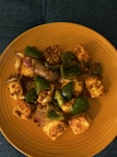 A dish featuring cubes of paneer seasoned with spices, mixed with sautéed green bell peppers and onion pieces, served on a round yellow plate. The presentation is simple and appetizing, with a focus on the vibrant colors and textures of the ingredients.