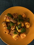 A dish featuring cubes of paneer seasoned with spices, mixed with sautéed green bell peppers and onion pieces, served on a round yellow plate. The presentation is simple and appetizing, with a focus on the vibrant colors and textures of the ingredients.