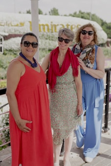 Three women are standing together, smiling and posing for a photo outdoors. They are dressed in colorful, summery outfits, with one woman in a red dress, another in a blue jumpsuit, and the third in a patterned dress with a red scarf. They all wear sunglasses, and the background features a hedge shaped like an airplane with the word 'Emirates' visible.