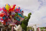 A person holding a large bundle of colorful balloons with various designs such as cartoon characters and animals. In the background, there is a large green topiary sculpture shaped like an elephant and a castle-like structure with some flowers. The sky is partly cloudy.