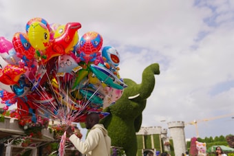 A person holding a large bundle of colorful balloons with various designs such as cartoon characters and animals. In the background, there is a large green topiary sculpture shaped like an elephant and a castle-like structure with some flowers. The sky is partly cloudy.