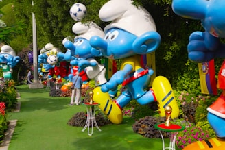 Large blue cartoon-like statues resembling animated characters are arranged in a garden setting, surrounded by greenery and flowers. The statues hold soccer balls and wear colorful sports gear. Bright flags and whimsical elements, such as miniature football trophies, add to the playful atmosphere.