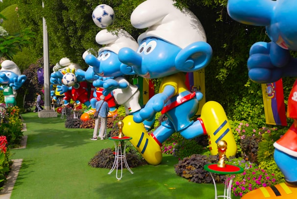 Large blue cartoon-like statues resembling animated characters are arranged in a garden setting, surrounded by greenery and flowers. The statues hold soccer balls and wear colorful sports gear. Bright flags and whimsical elements, such as miniature football trophies, add to the playful atmosphere.