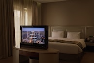 A cozy hotel room with a neatly made bed featuring white pillows and a beige bedspread. A modern flat-screen television is placed on an elegant table in front of the bed, displaying a welcome message from a place called Tower Plaza. The room is softly lit with natural light coming from a window with long curtains drawn to the side. The neutral color palette and minimalist design create a calm and relaxing atmosphere.