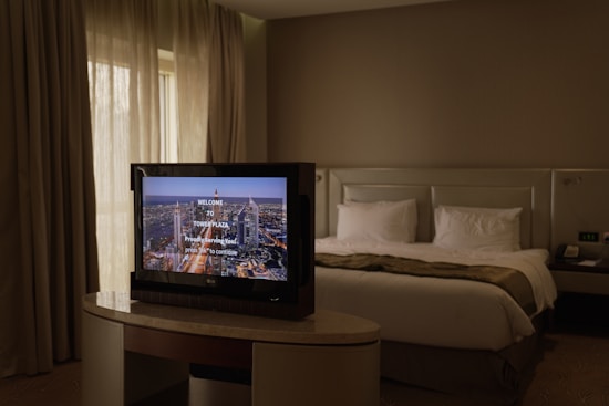 A cozy hotel room with a neatly made bed featuring white pillows and a beige bedspread. A modern flat-screen television is placed on an elegant table in front of the bed, displaying a welcome message from a place called Tower Plaza. The room is softly lit with natural light coming from a window with long curtains drawn to the side. The neutral color palette and minimalist design create a calm and relaxing atmosphere.