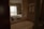 A modern bathroom with a white freestanding bathtub in the foreground. Above the bathtub, there are two white towels neatly rolled and hanging on a rack. The room is softly lit with a window in the background showcasing tall urban buildings partially visible from behind a lightly drawn curtain. A neatly made bed with a green accent runner rests just beyond a set of wooden louvered doors that partially separate the room from the bed view.