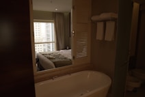 A modern bathroom with a white freestanding bathtub in the foreground. Above the bathtub, there are two white towels neatly rolled and hanging on a rack. The room is softly lit with a window in the background showcasing tall urban buildings partially visible from behind a lightly drawn curtain. A neatly made bed with a green accent runner rests just beyond a set of wooden louvered doors that partially separate the room from the bed view.