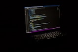 A laptop with an illuminated keyboard displays lines of code on its screen against a dark background. The code is highlighted with various colors to indicate syntax.